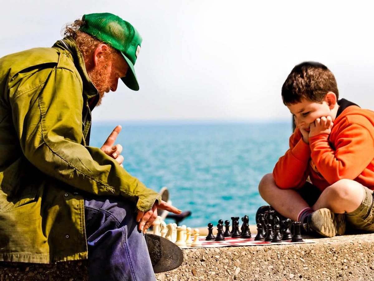 An adult helps a child think through a move on a chessboard by the lake.